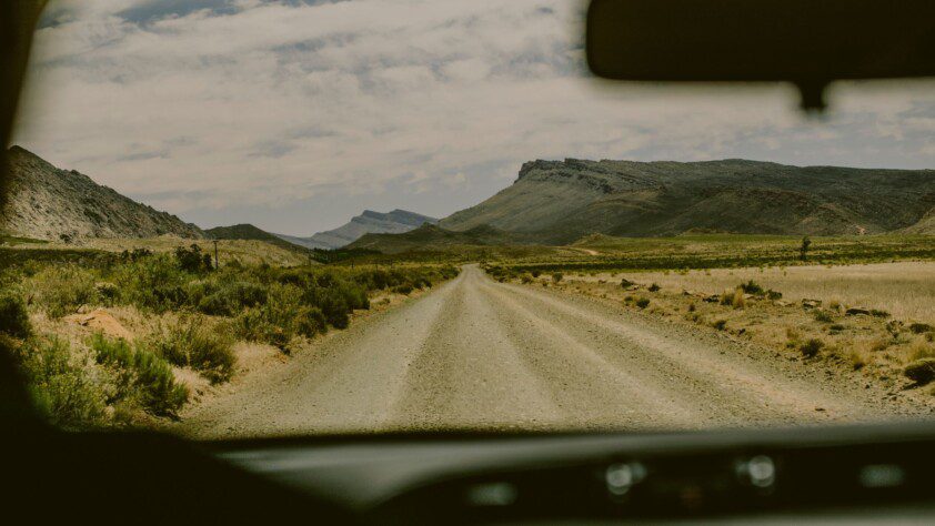 View from a vehicle's interior of a gravel road leading through the rugged terrain of Southern Africa, under a vast sky.