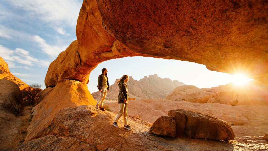 Two hikers who have used 4x4 rentals to reach a majestic rock arch at sunset in Namibia.
