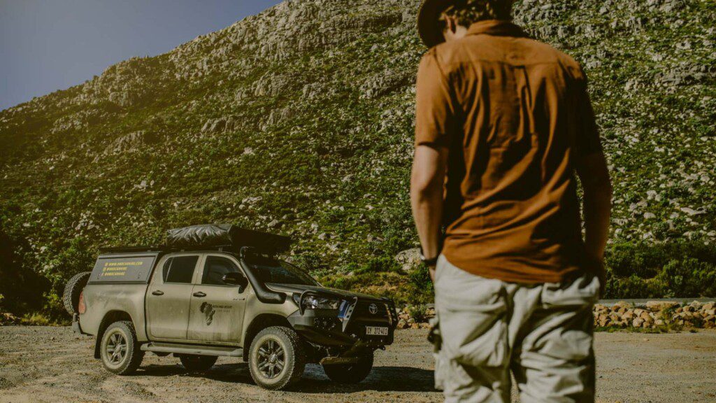 Adventurer standing near a rugged Toyota Hilux 4WD in a mountainous area, ready for an overlanding journey.
