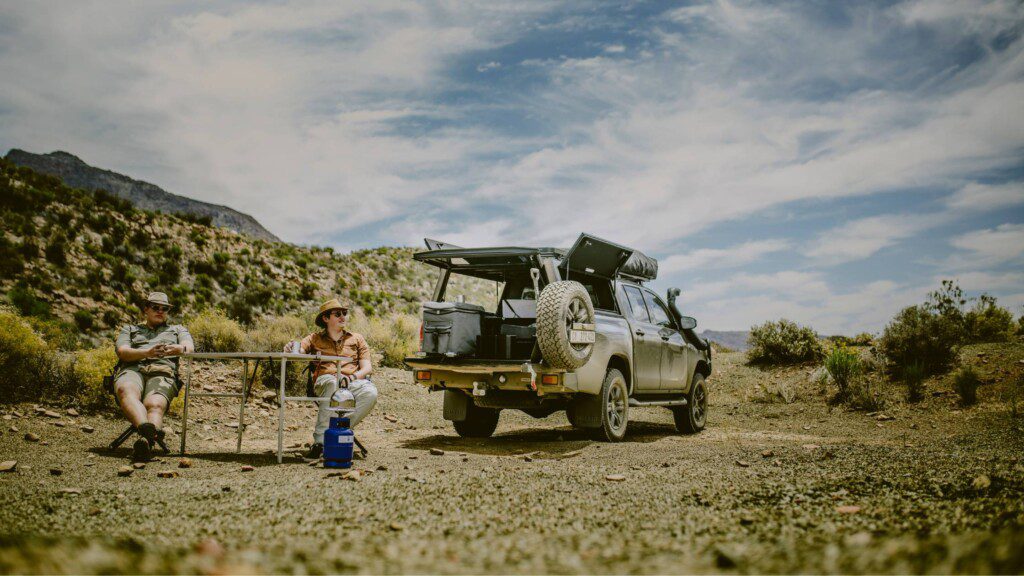 Bush and Desert Car hire: Two travellers relaxing next to a fully equipped 4x4 vehicle in a scenic desert landscape.