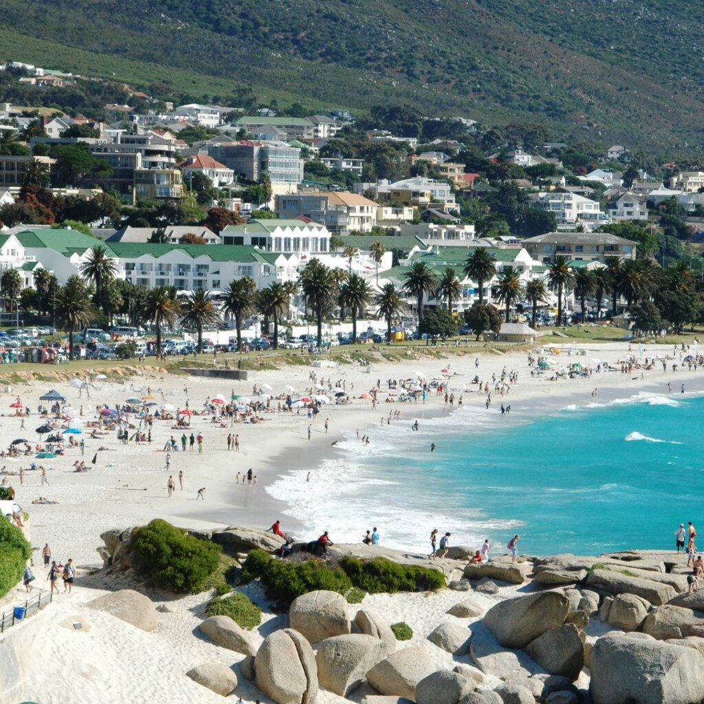 A lively scene at Camps Bay Beach in Cape Town, South Africa, showcasing a bustling beachfront with people enjoying the sun, sea, and sand.